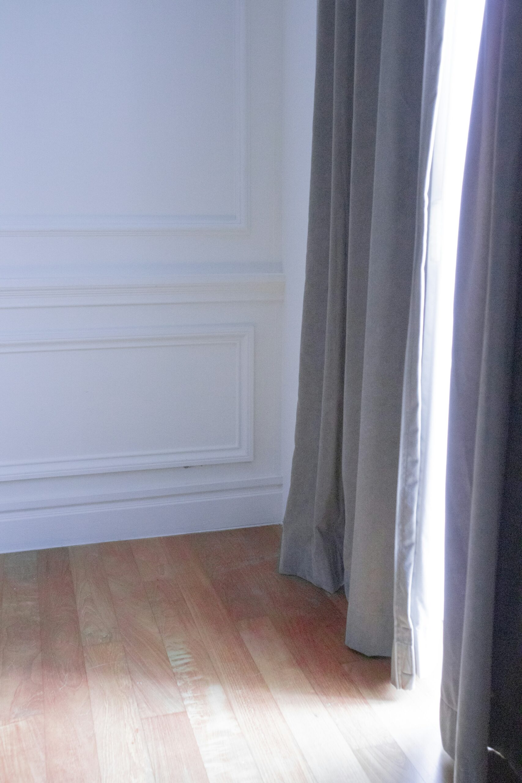 A corner of a room with light-colored walls, wooden floor, and dark curtains partially open to let in natural light.