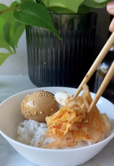 A bowl of white rice topped with a marinated egg and kimchi is being served with chopsticks; a green plant is in the background.