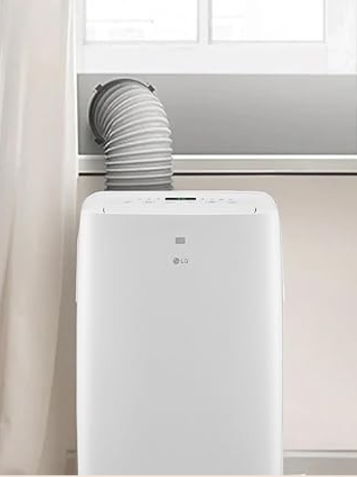 A portable air conditioner is positioned near a window with an exhaust hose vented outside. Nearby, a massage gun rests on the table beside a curtain and a potted plant, creating a comfortable and relaxing environment.