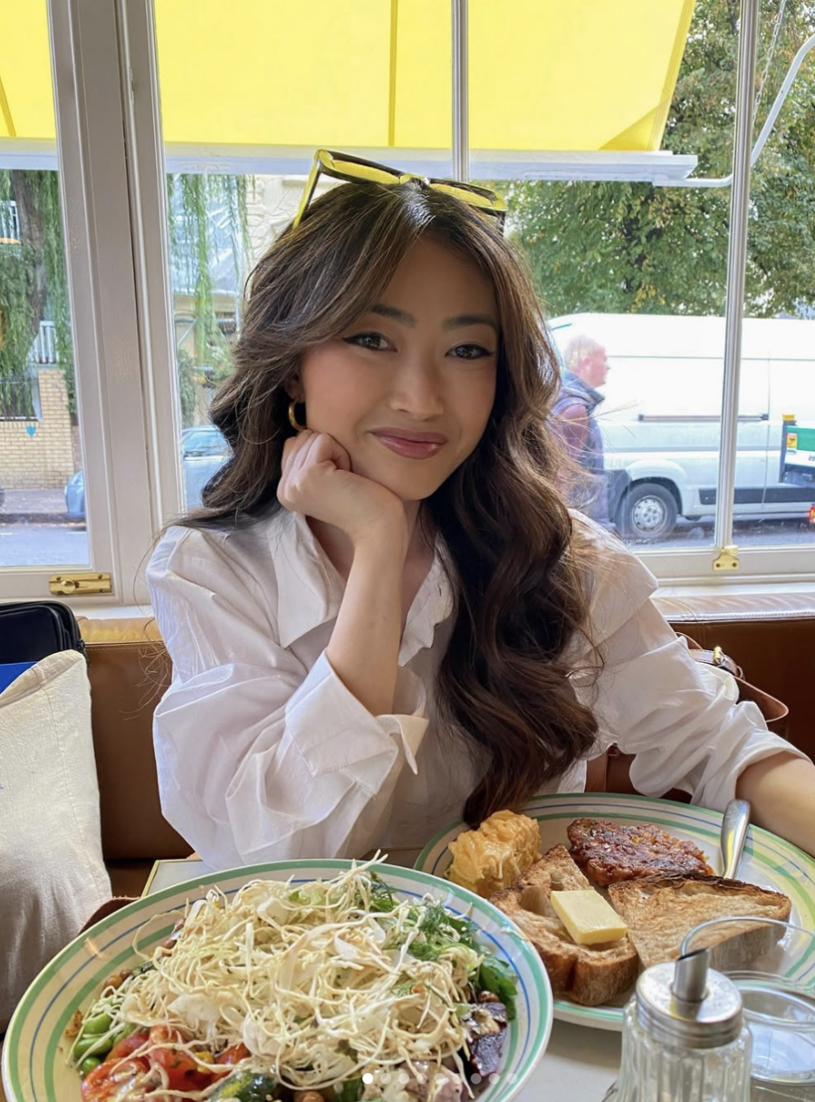 A woman in a white shirt sits at a table in a cafe with plates of salad, bread, and other food in front of her, smiling at the camera.