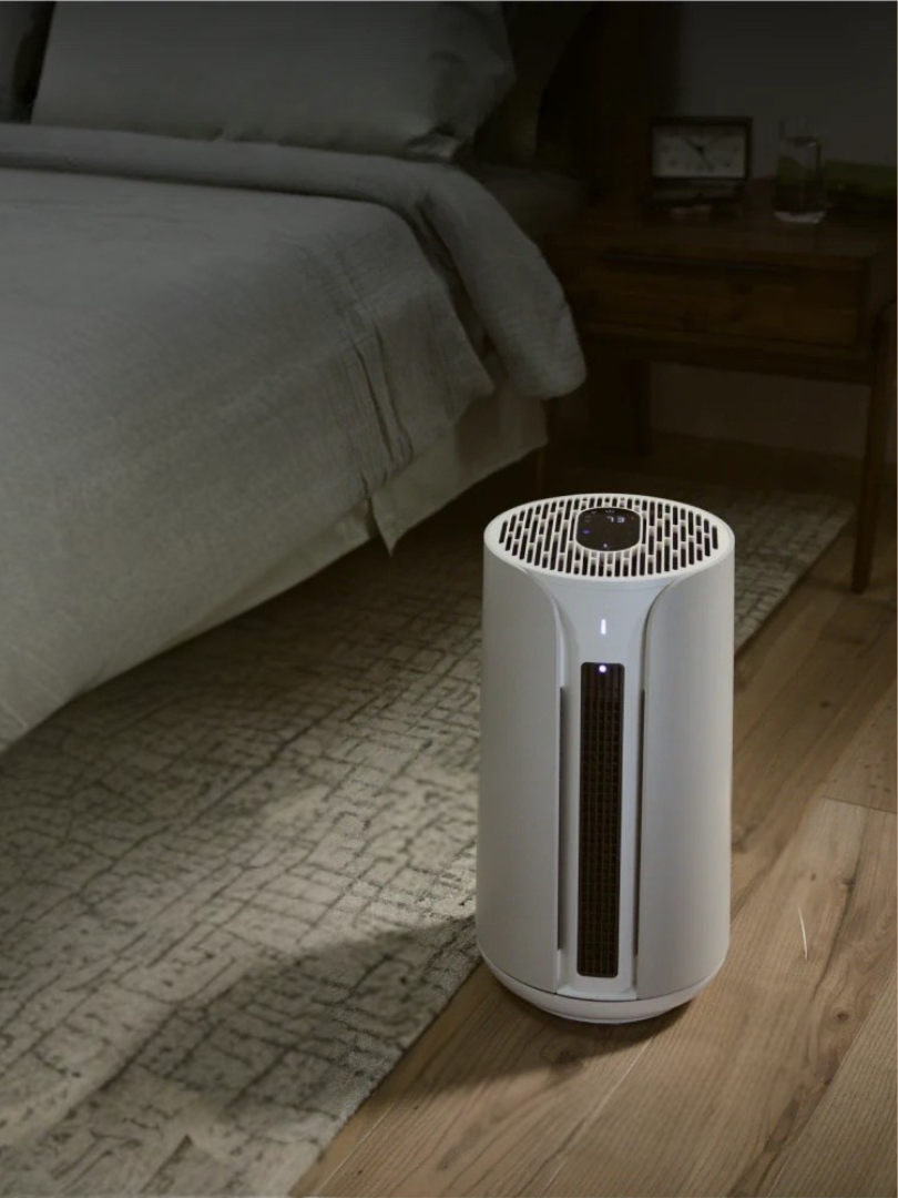 A white air purifier sits on a wooden floor next to a bed in a dimly lit bedroom, with massage guns neatly stored nearby for relaxation and comfort.
