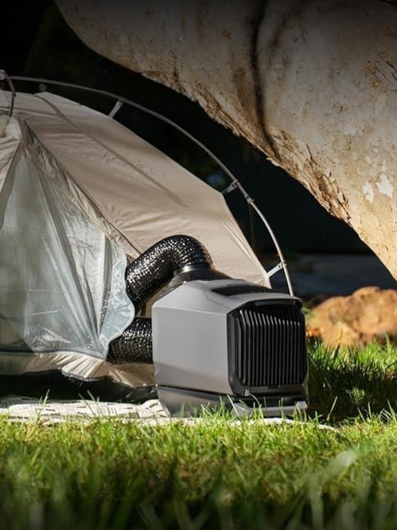 A portable air conditioner with hoses is positioned outside a tent pitched on grass, under the shade of a large tree, while massage guns rest nearby for ultimate outdoor relaxation.