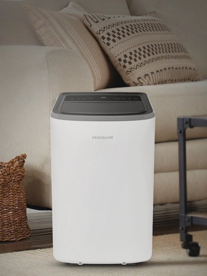 A white Frigidaire portable air conditioner is positioned in front of a beige sofa in a living room setting, creating a relaxing space perfect for unwinding with massage guns after a long day.