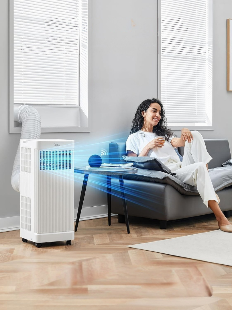 A woman sits on a sofa holding a mug, while a portable air conditioner and massage guns rest nearby, keeping her comfortable in a bright, modern room.