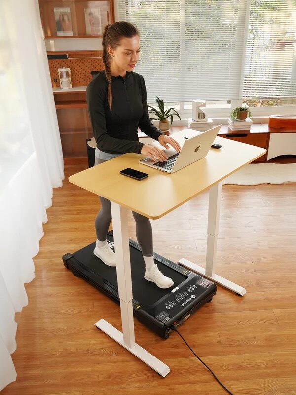 A woman uses a laptop on a standing desk while walking on a treadmill in a bright home office with wooden floors.