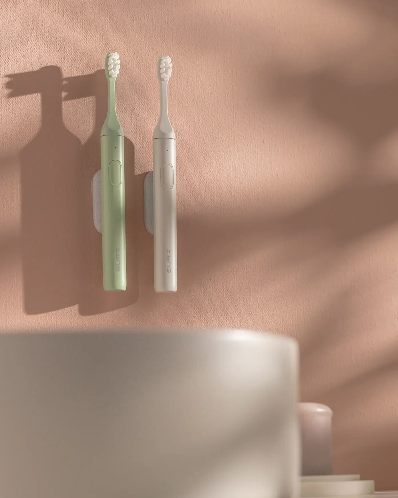 Two electric toothbrushes, one green and one white, are mounted vertically on a pink wall with soft shadows cast across the scene.