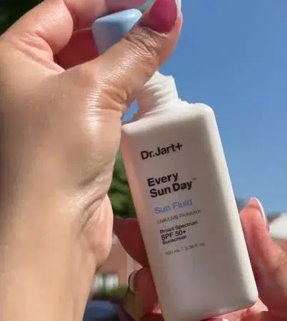 A hand holds a bottle of Dr. Jart+ Every Sun Day Sun Fluid SPF 50+ outdoors under a clear blue sky.