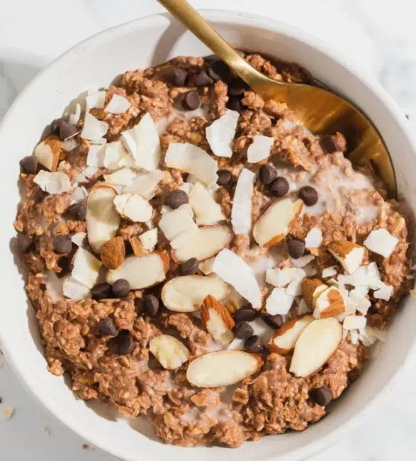 A bowl of chocolate oatmeal topped with almond slices, coconut flakes, and chocolate chips, with a gold spoon resting in the bowl.