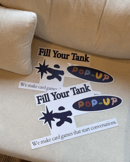 Stickers on a beige couch, including “Fill Your Tank,” “POP-UP,” and one reading, “We make card games that start conversations.”.