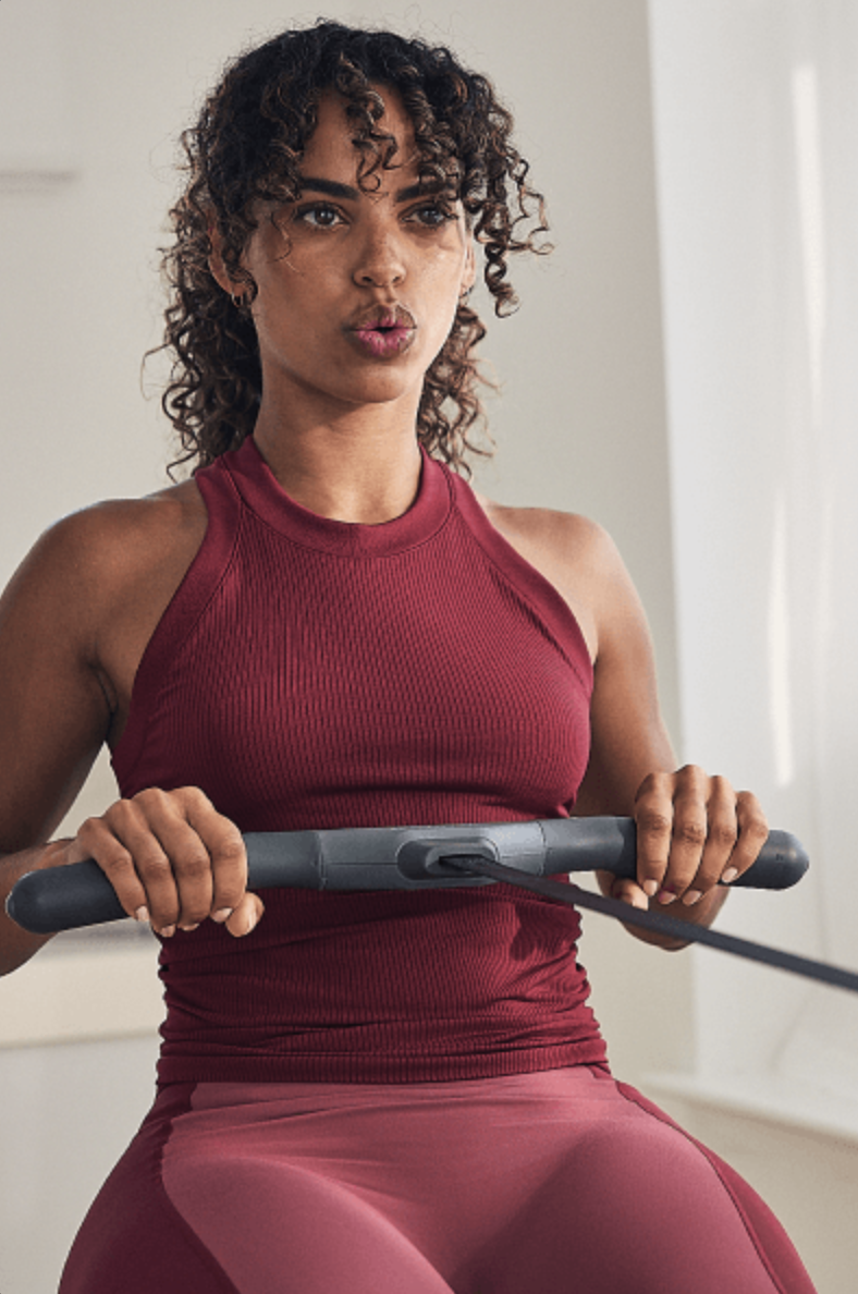 A woman in a burgundy athletic outfit exercises on a rowing machine indoors, looking focused—perfectly pairing her workout with massage guns for optimal recovery.