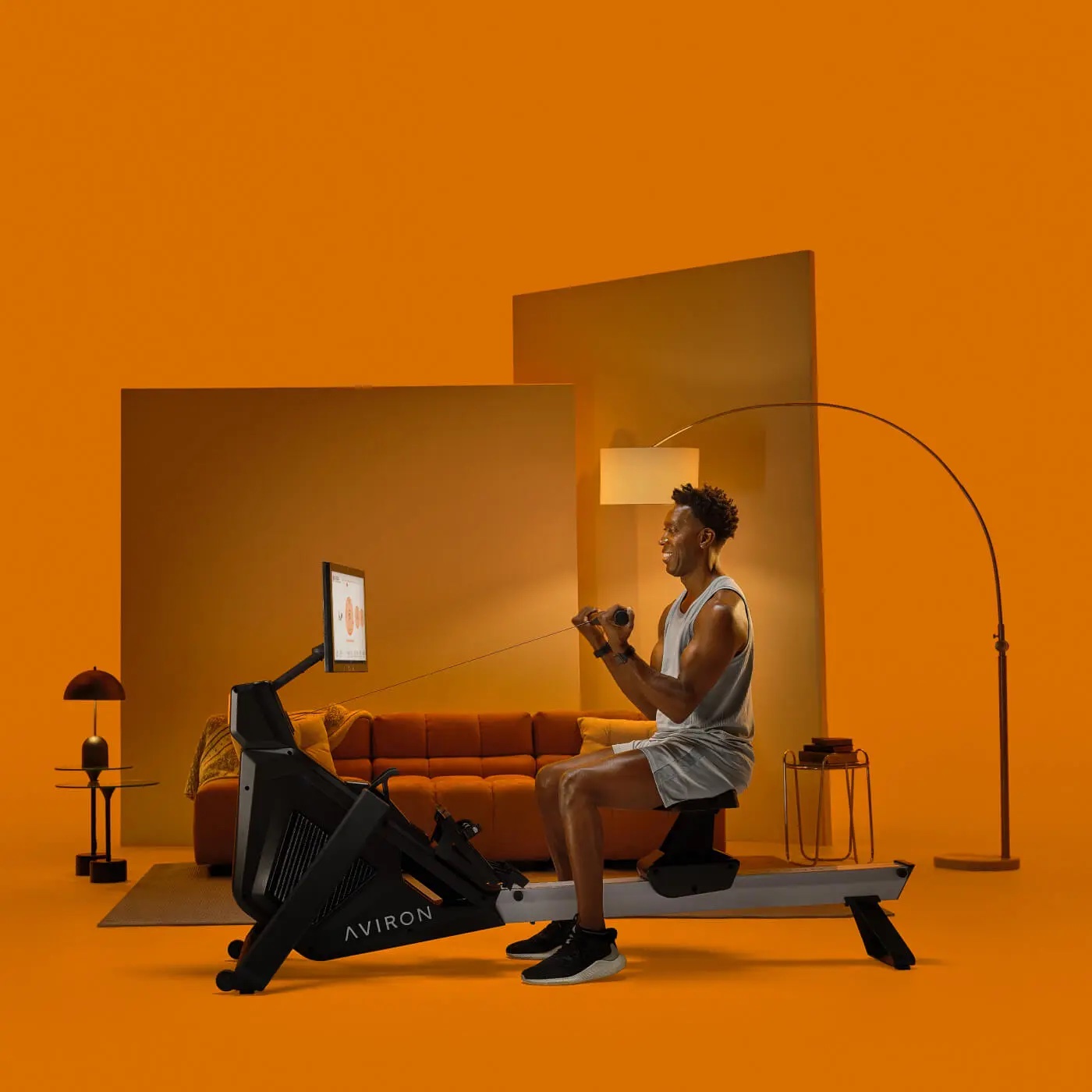 A person exercises on an Aviron rowing machine in a modern, orange-themed living room with a sofa, lamp, decor, and massage guns visible in the background.