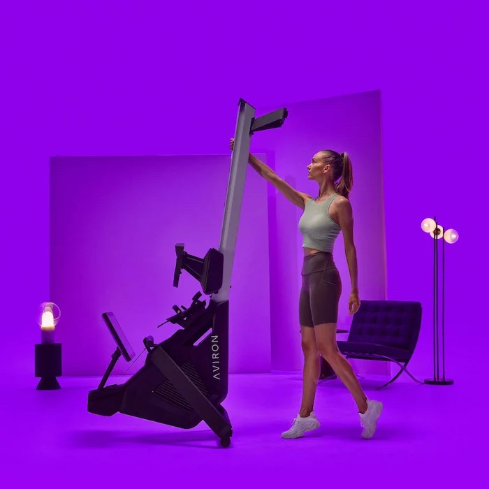 A woman in workout clothes stands in a purple-lit room, reaching up to adjust a vertical rowing machine labeled "Aviron," while massage guns rest nearby for post-workout recovery.