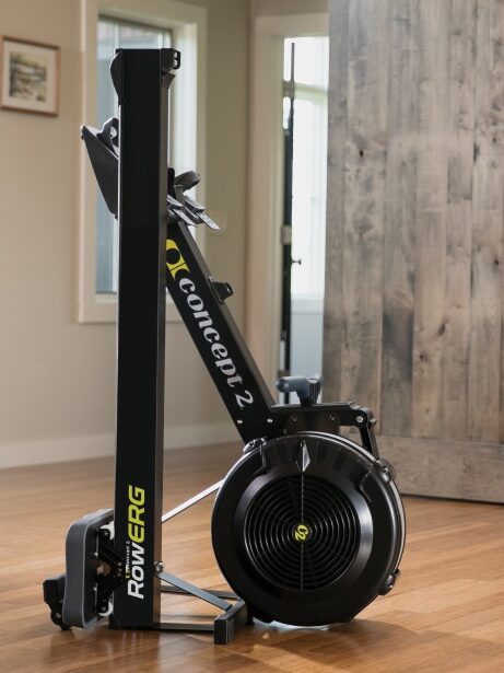 A Concept2 RowErg rowing machine sits on a wooden floor in a modern, well-lit room with massage guns nearby, a sliding barn door, and framed pictures adorning the walls.