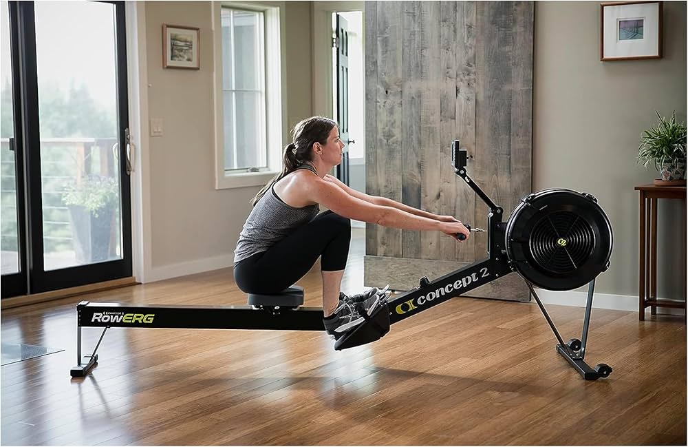 A person exercises indoors on a Concept2 rowing machine, positioned in a well-lit room with large windows and wooden flooring, keeping massage guns nearby for post-workout muscle recovery.