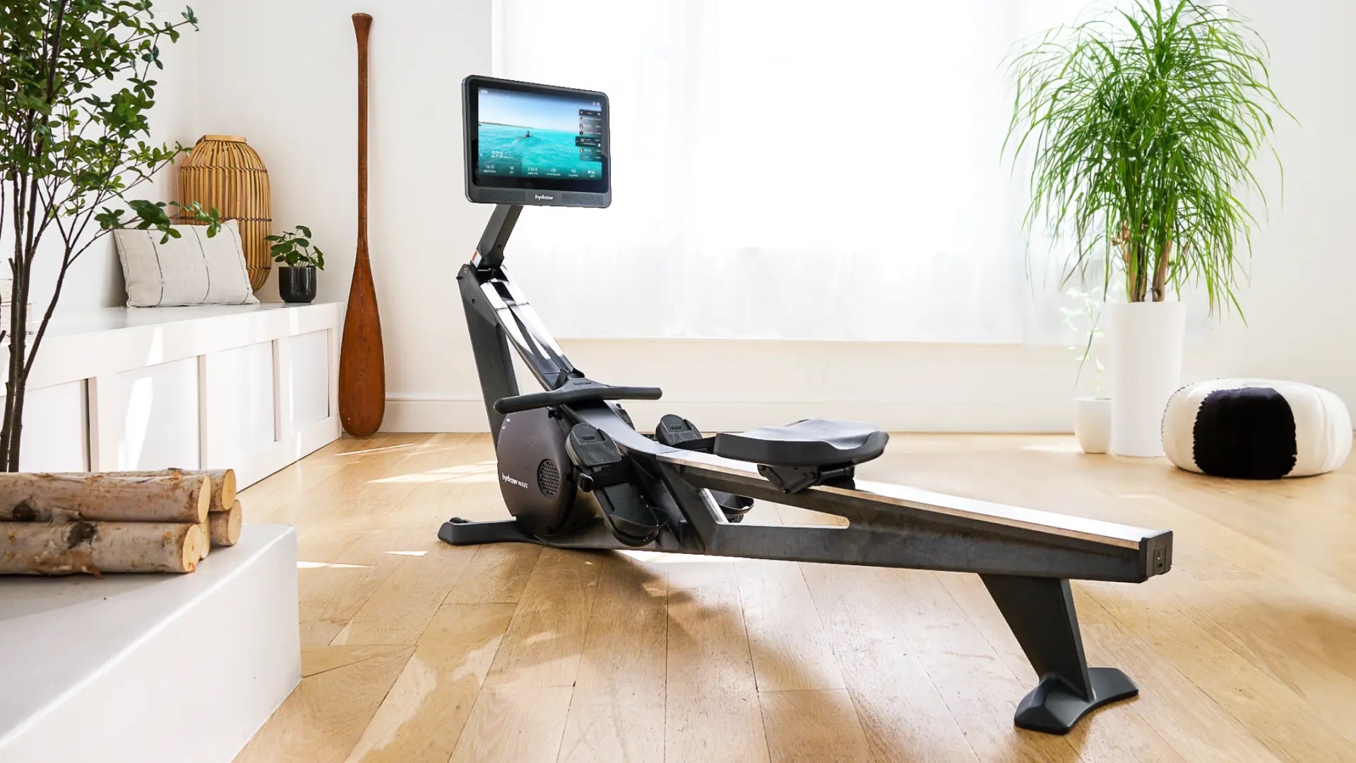 A modern rowing machine with a large screen sits in a bright, minimalist room with wooden floors, plants, and natural light from a window—perfectly paired with massage guns for post-workout recovery.