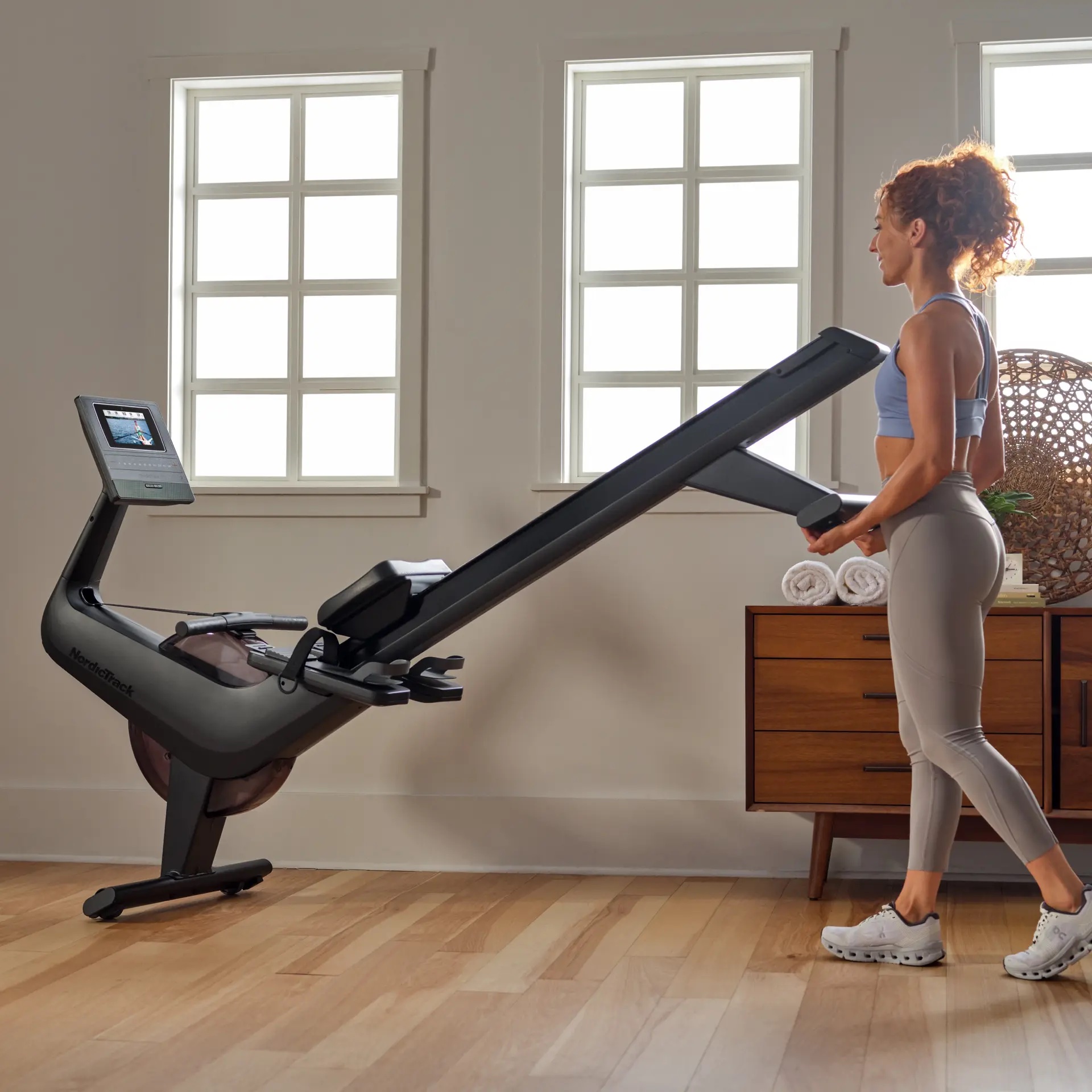 A woman in workout clothes folds up a rowing machine in a bright, modern room with wooden floors, a dresser in the background, and massage guns neatly arranged on top.