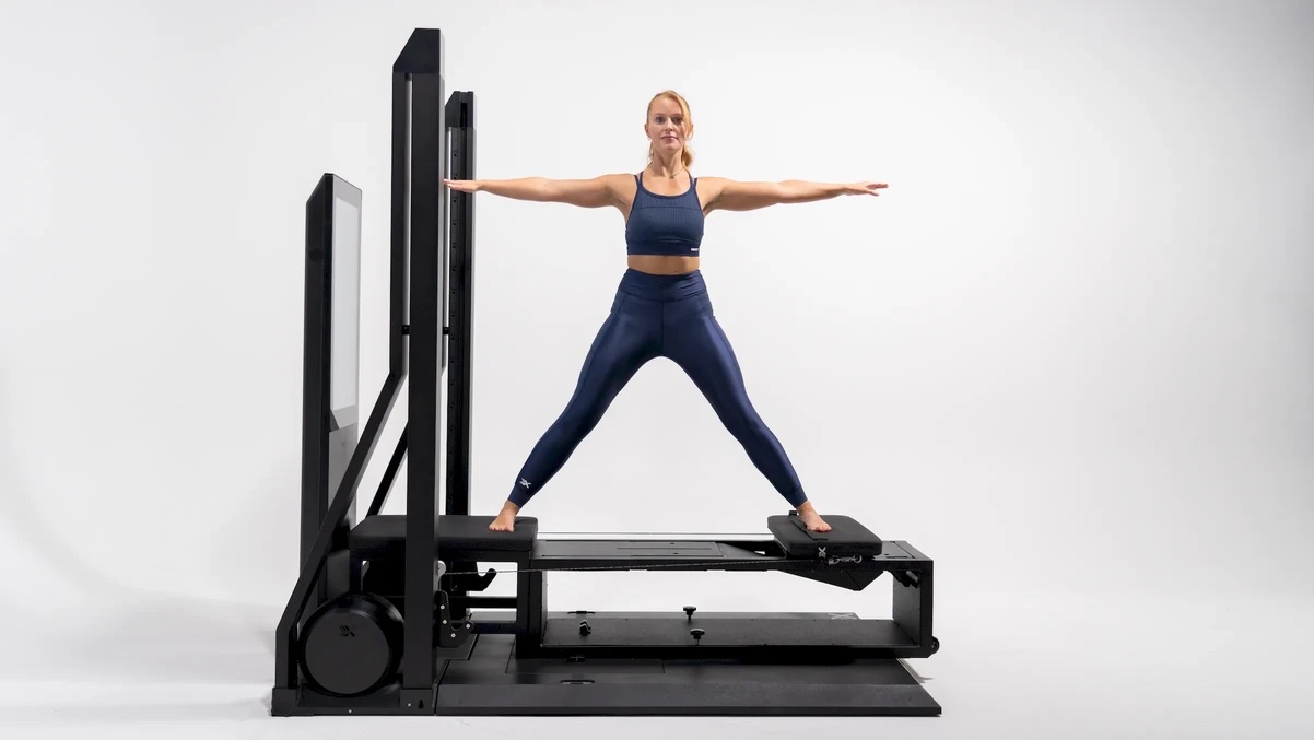 A woman in athletic wear stands in a wide-leg stance with arms outstretched on a black fitness reformer machine in a white studio, where massage guns are often used for post-workout recovery.