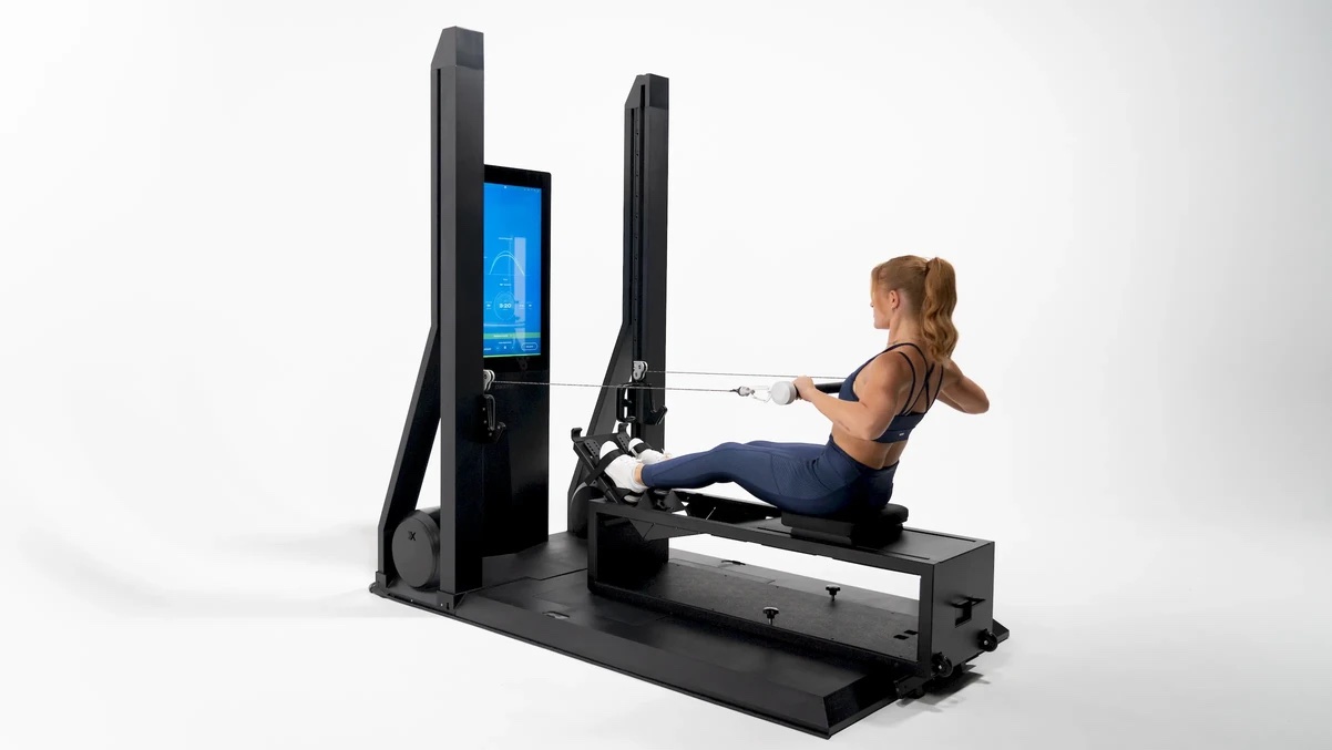 A woman uses a modern rowing machine with a large built-in digital screen against a plain white background, ideal for pairing with massage guns to aid in post-workout recovery.