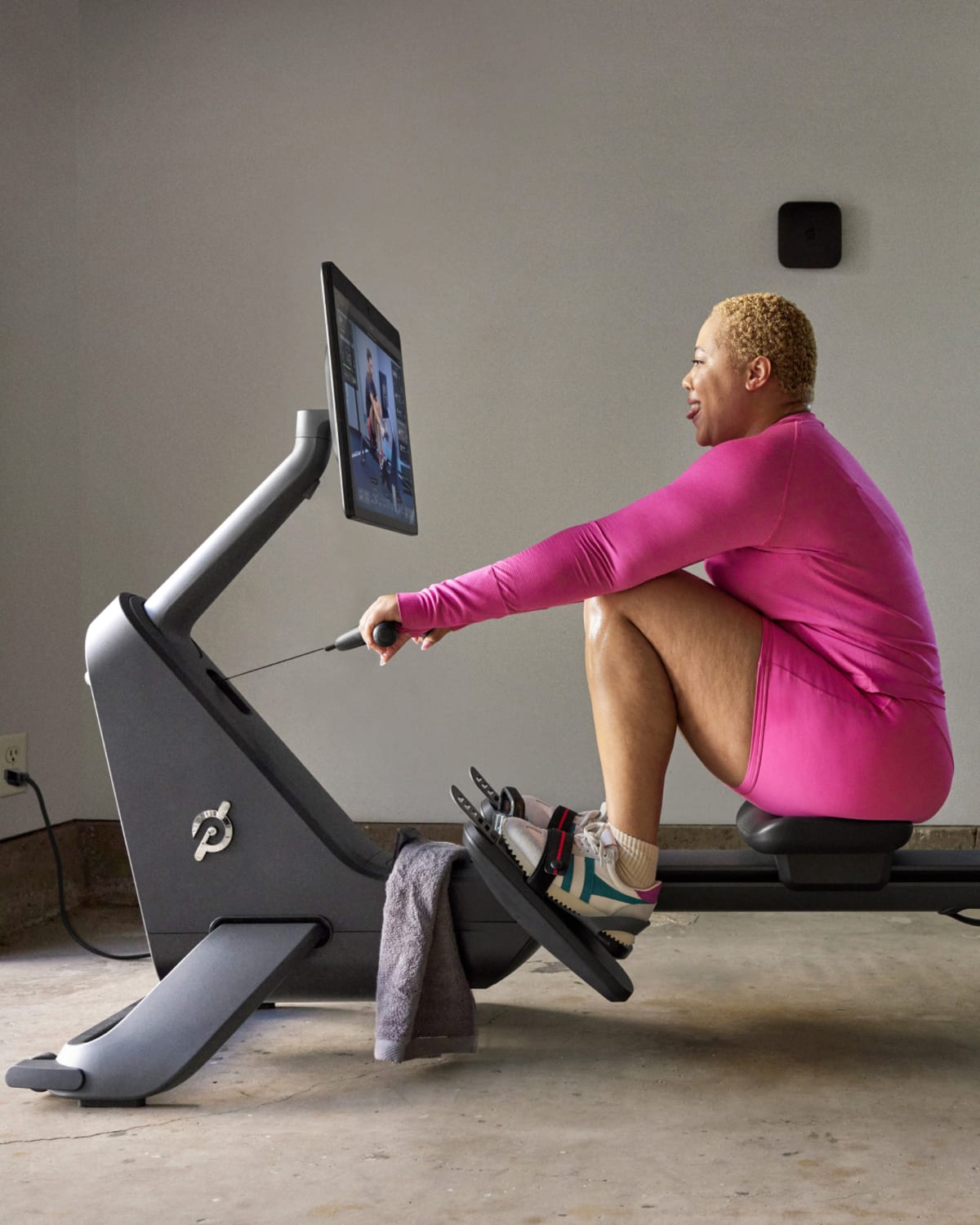 A person wearing a bright pink outfit uses a rowing machine while watching a workout on a screen in a home gym, with massage guns nearby for post-exercise recovery.