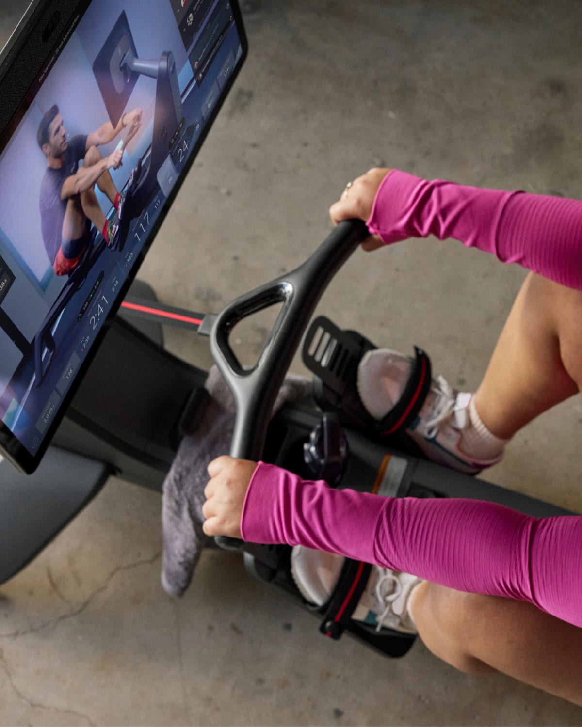 Person wearing pink sleeves uses a stationary bike while watching a virtual cycling instructor on a screen, with massage guns visible nearby for post-workout recovery.