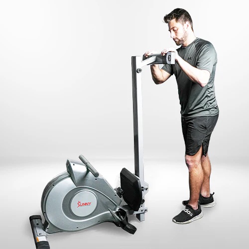A man in athletic wear stands next to a Sunny rowing machine, adjusting the upright rail in a well-lit studio, while nearby, massage guns rest on a bench, highlighting recovery tools for his workout.