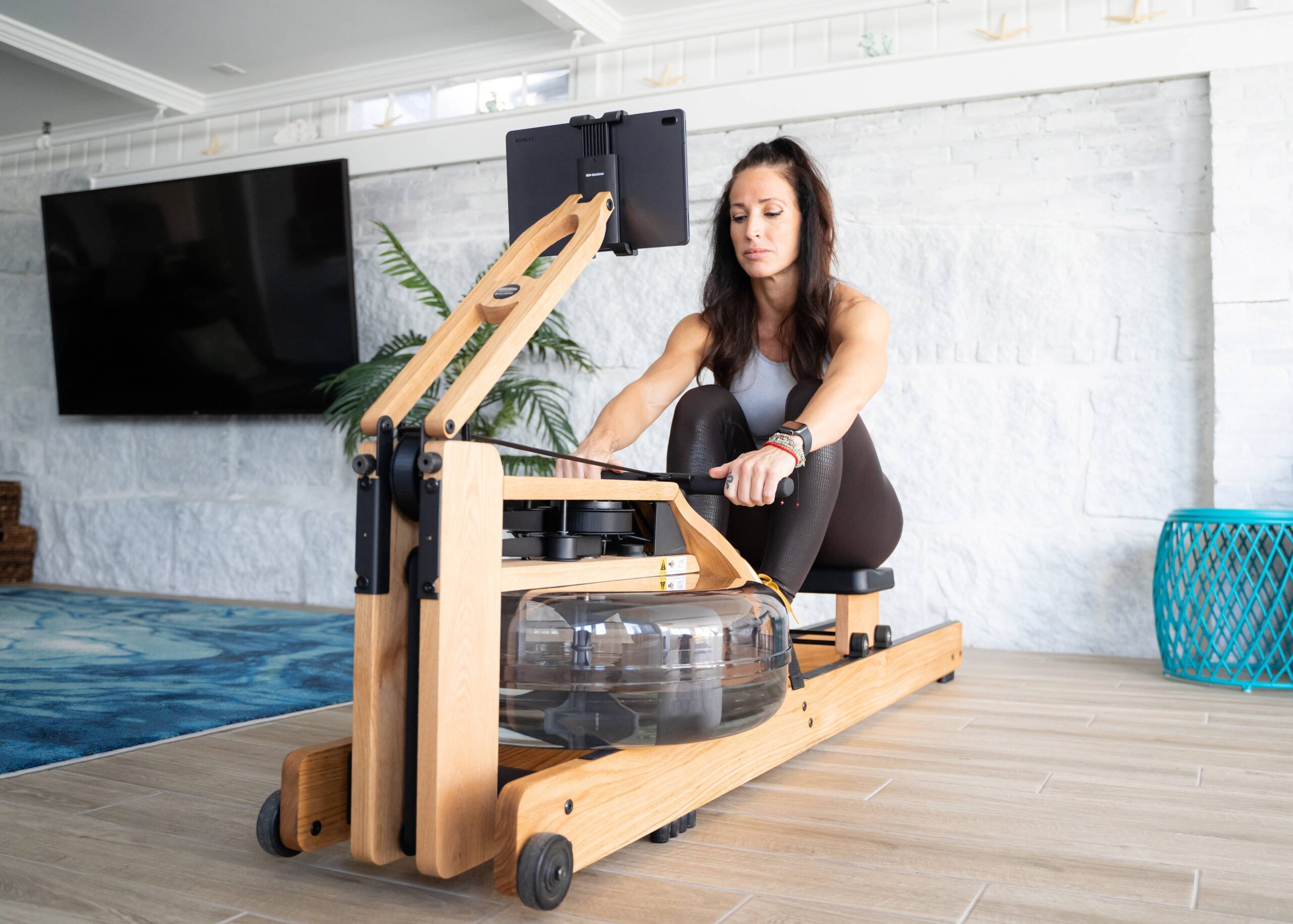 Woman sitting on a wooden rowing machine indoors, adjusting the foot straps while facing a monitor attached to the equipment. Massage guns rest nearby, ready to help soothe muscles after her workout.
