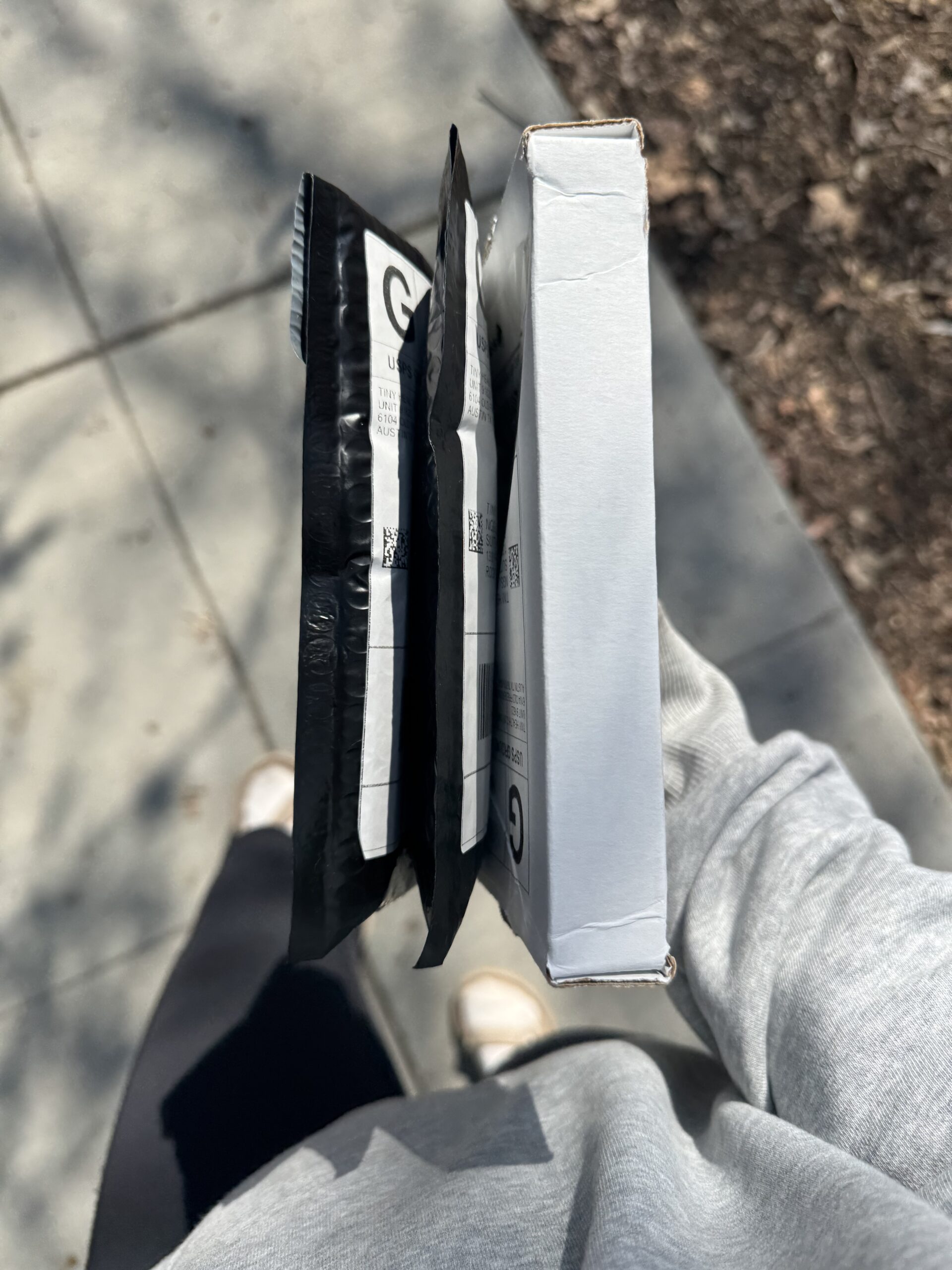 A person in a gray sweatshirt holds two black padded envelopes and one white box while standing on a sidewalk.