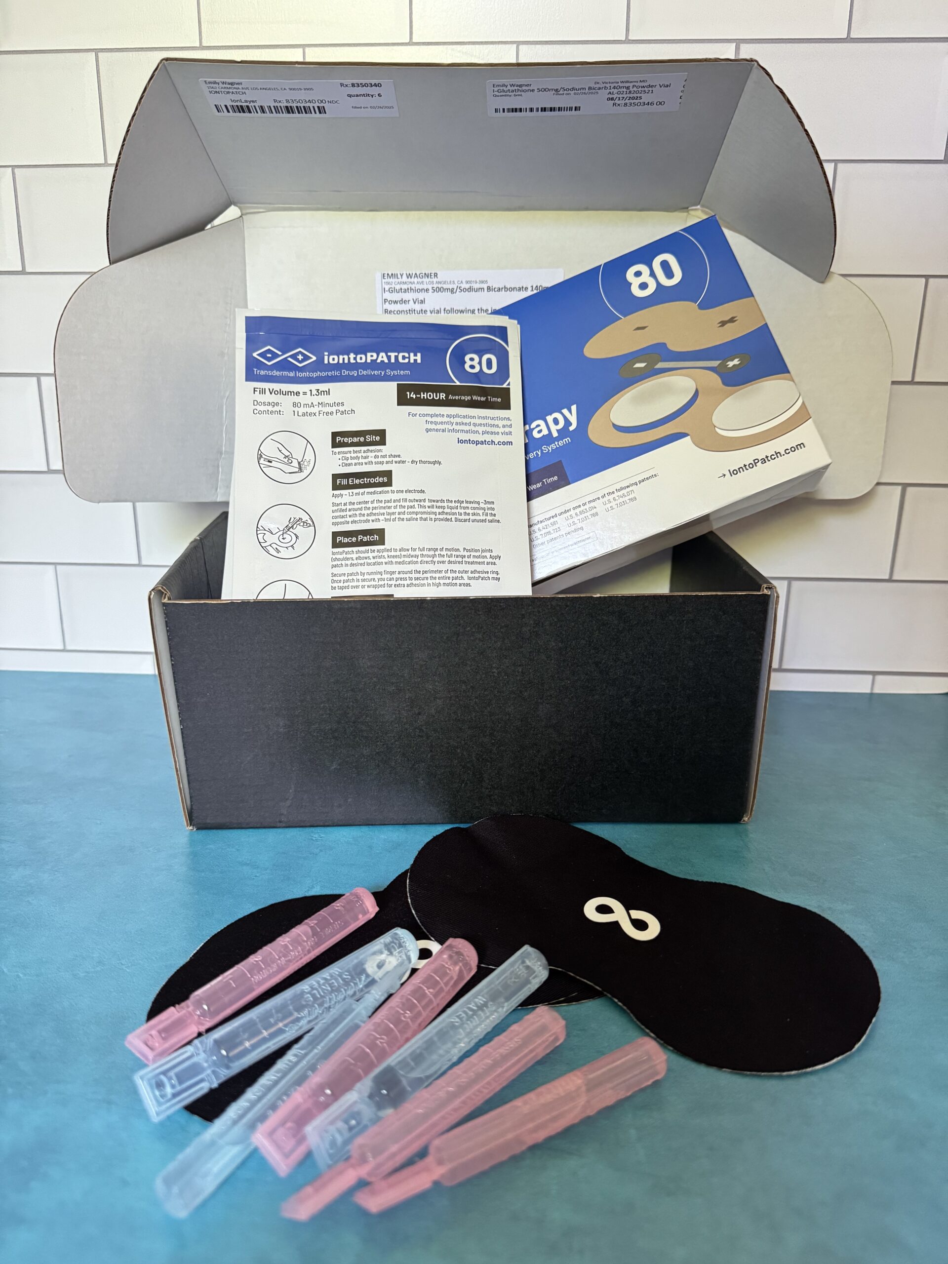 A black box with IontePATCH therapy supplies including instruction sheets, adhesive patches, two black eye masks, and several pink and blue plastic clips on a blue surface.