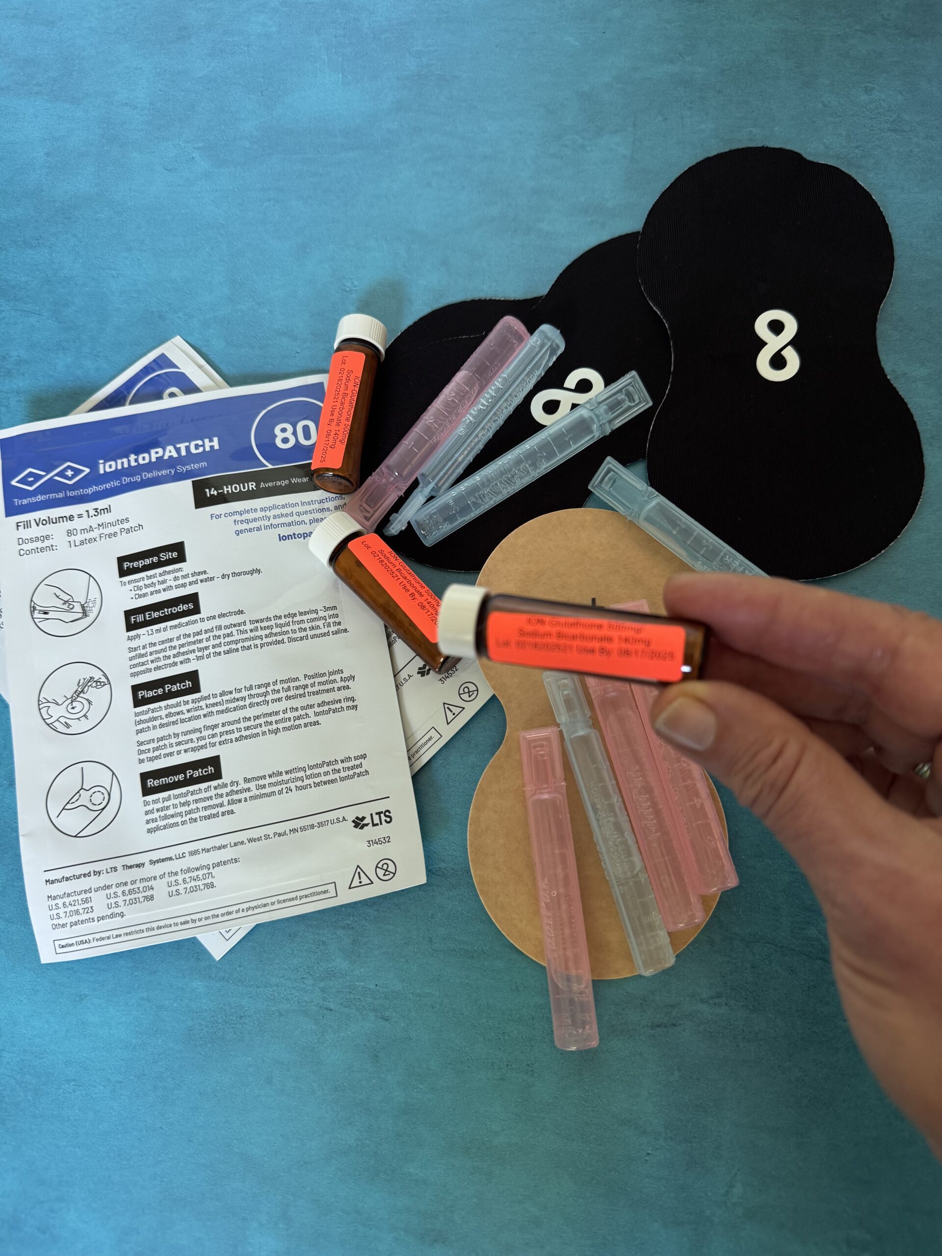 A hand holds a labeled tube over a table with iontoPATCH instructions, multiple small tubes, two black patches with the number 8, and a round adhesive pad on a blue surface.