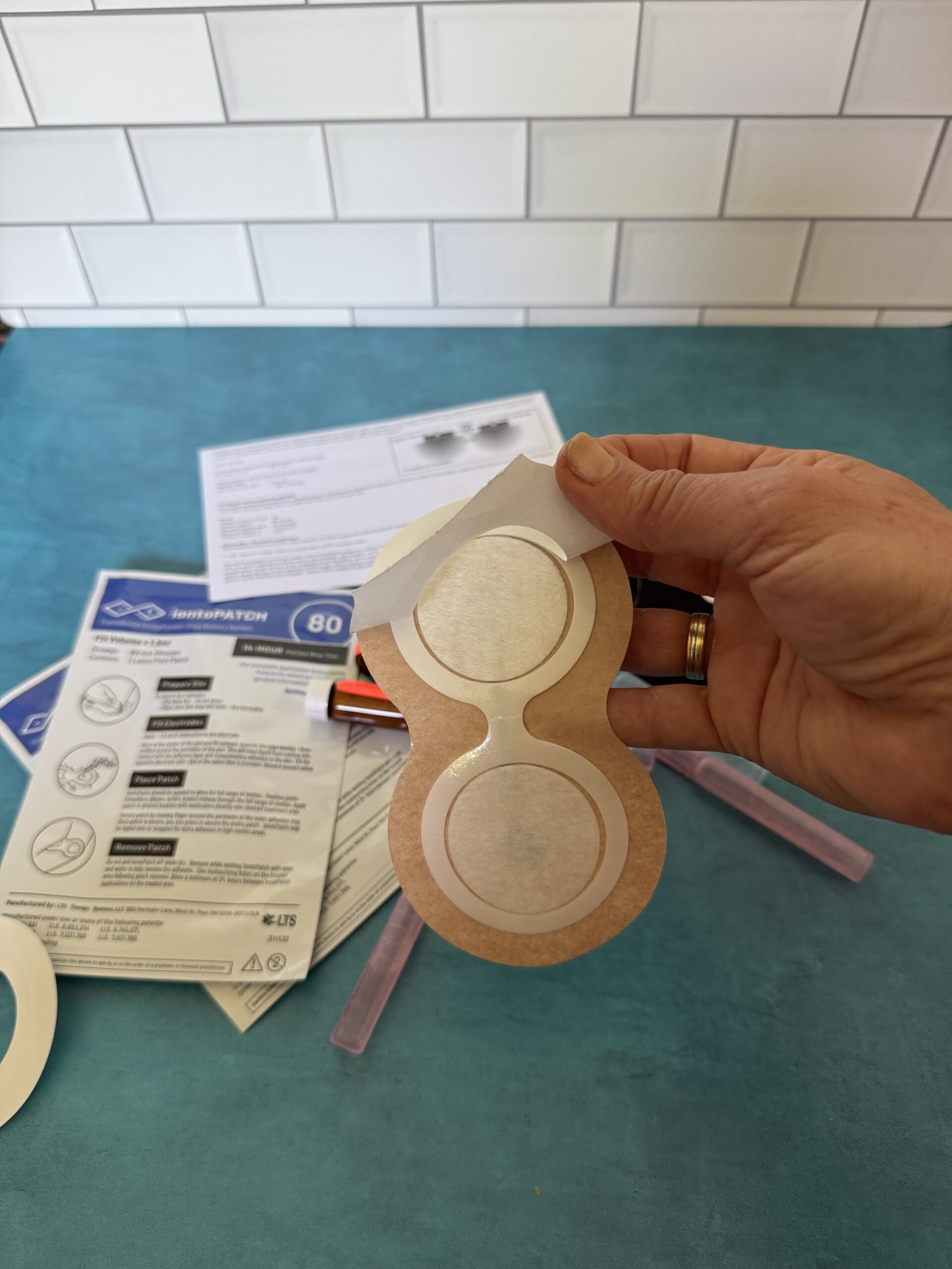 A hand holds a medical adhesive patch with backing partially peeled off; instruction sheets and other patches are on a blue surface in the background.