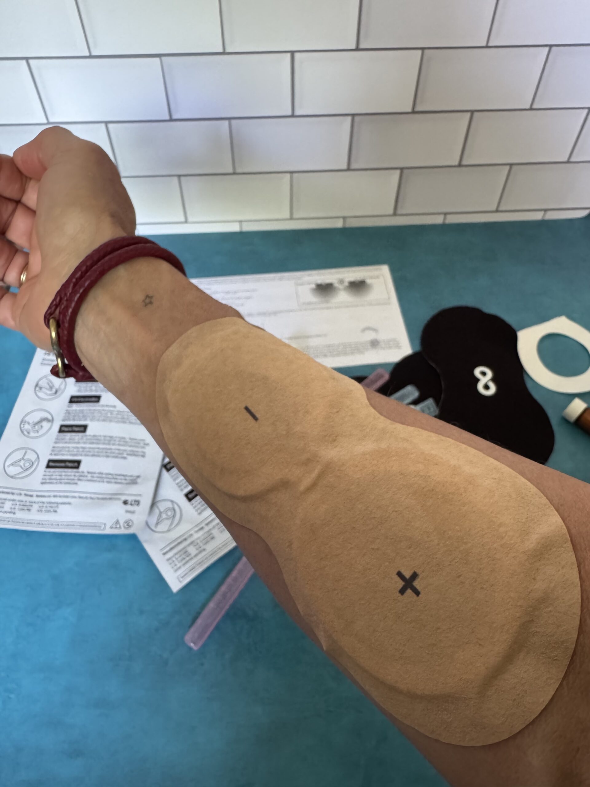 A forearm with large adhesive patches marked with "+" and "O", with instructions and medical supplies on a blue counter in the background.