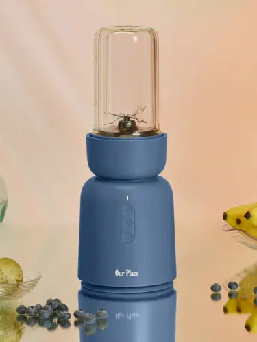 Blue portable blender with a transparent blending cup on top, displayed upright on a reflective surface alongside massage guns, blueberries, and a banana.