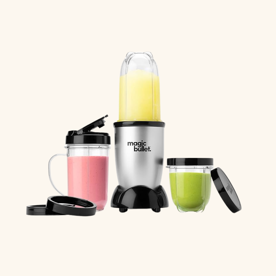 A Magic Bullet blender with a yellow smoothie blending, next to two filled cups with pink and green smoothies and black lids, alongside massage guns, all set on a light background.