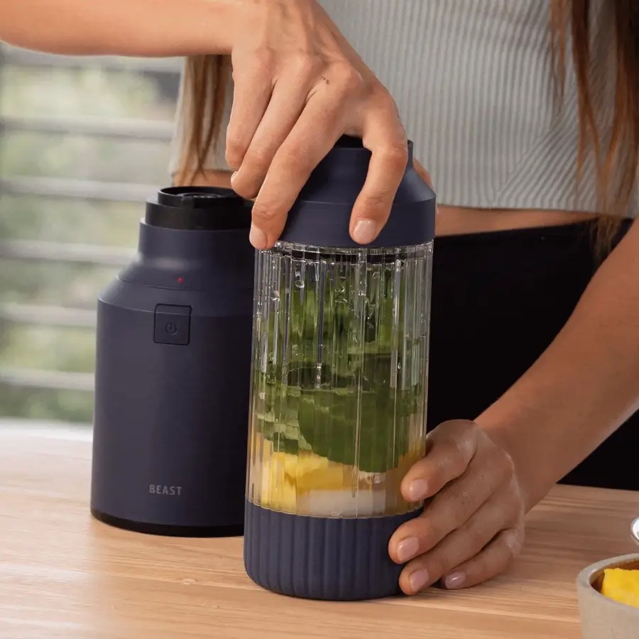 A person holds a portable blender filled with pineapple, spinach, and ice on a wooden table, preparing to blend the ingredients after an intense workout using massage guns.