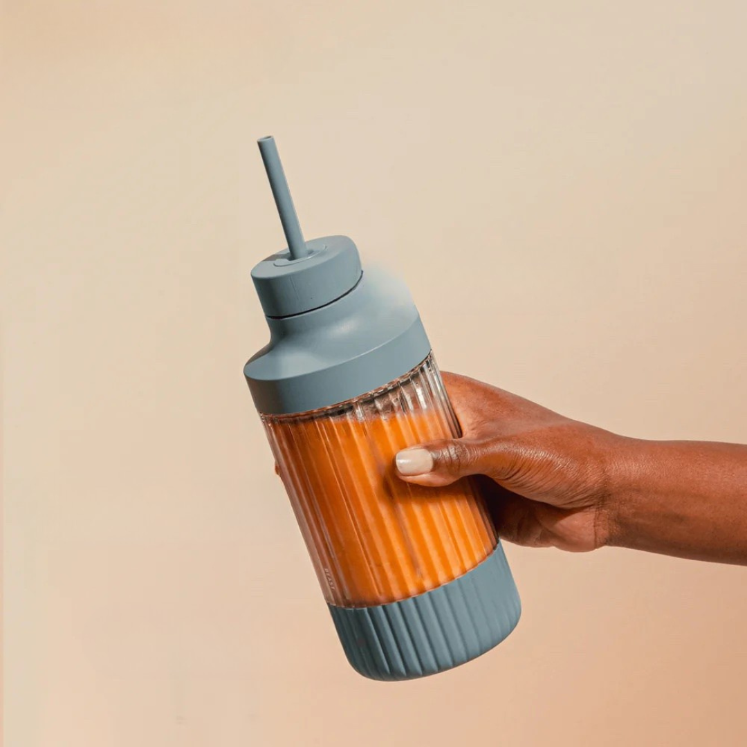 A hand holding a ribbed glass bottle with a blue lid and straw, filled with an orange beverage, sits against a neutral background—perfect refreshment after using massage guns.