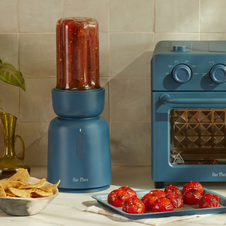 A blue blender with salsa, a blue toaster oven, a plate of roasted tomatoes, massage guns, and a bowl of tortilla chips sit on the kitchen counter.