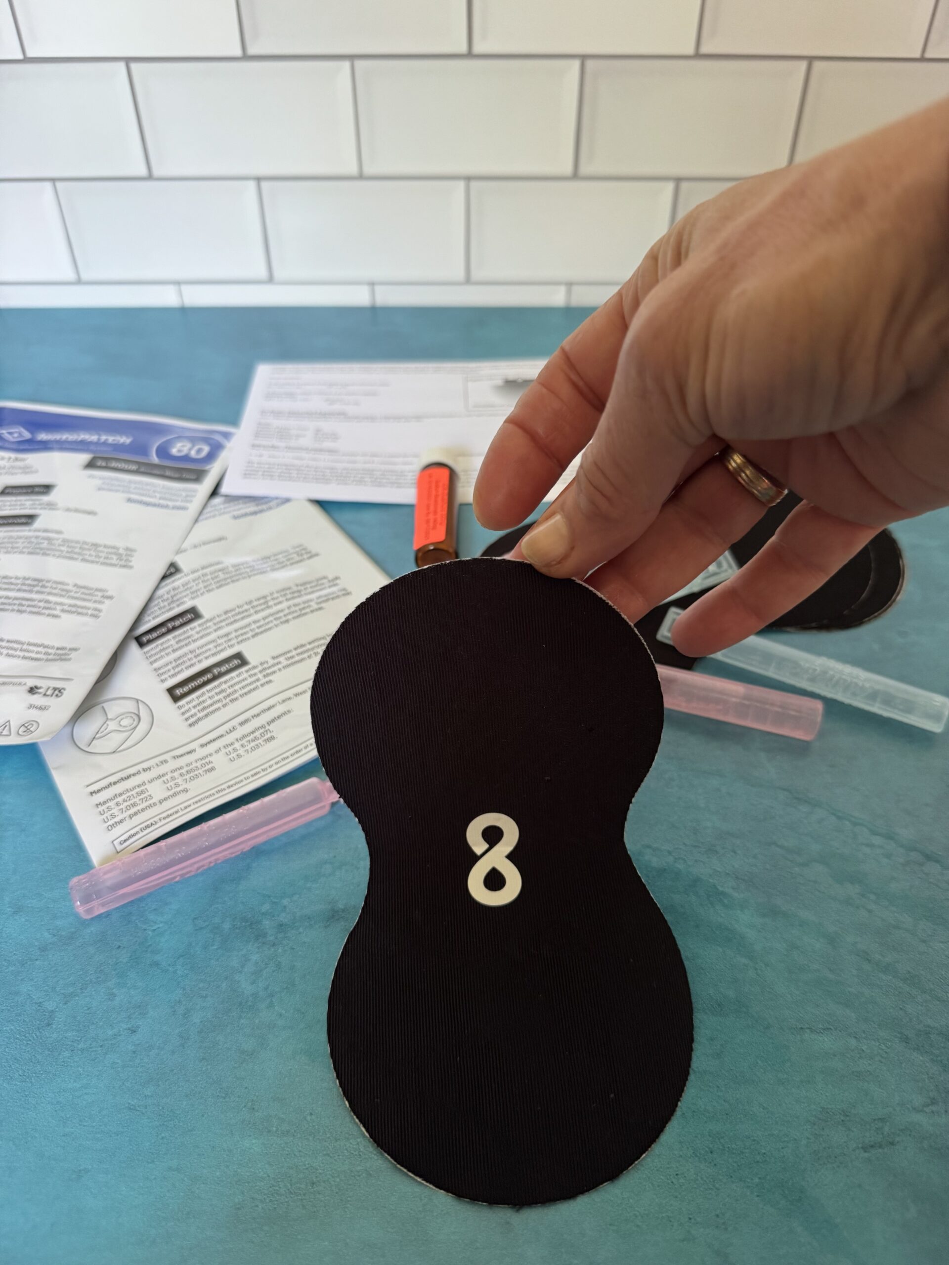 A hand holds a black figure-eight patch with the number 8 on it, above a blue surface with instructions and colored markers in the background.