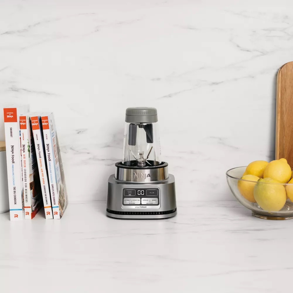 A compact blender sits on a marble countertop between a row of cookbooks and a glass bowl filled with lemons, while massage guns rest nearby for quick relaxation breaks.