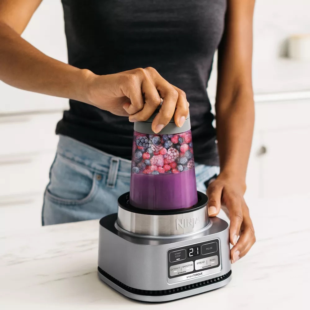 A person presses a blending cup filled with berries and liquid onto a Ninja blender base on a kitchen counter, while massage guns rest nearby.