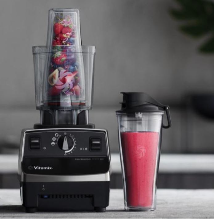 A Vitamix blender filled with various fruits sits on the countertop next to a lidded cup of pink smoothie—perfect fuel for a post-workout recovery session with massage guns.
