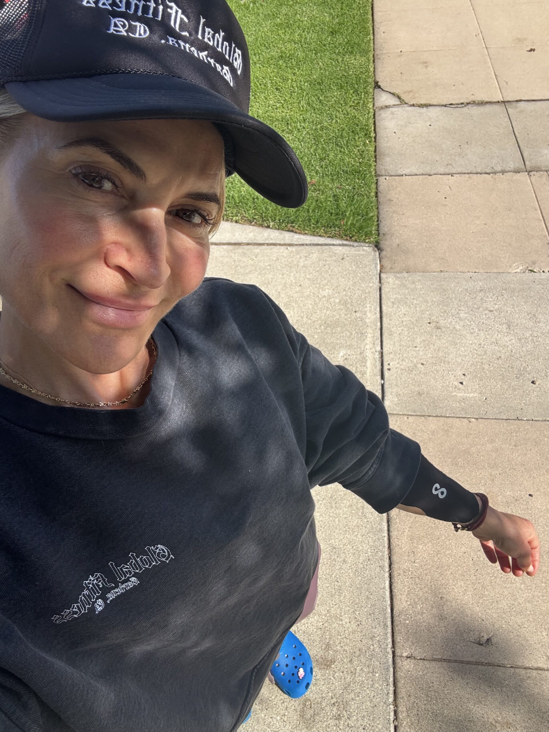 A person wearing a black sweatshirt, black cap, and blue Crocs stands on a sidewalk next to grass, taking a selfie outdoors.