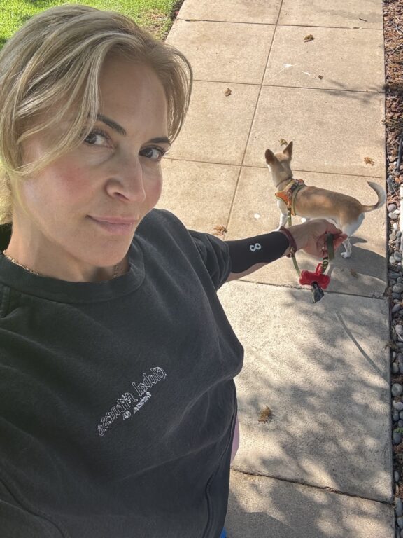 Woman taking a selfie outdoors while holding the leash of a small dog standing on a sidewalk.