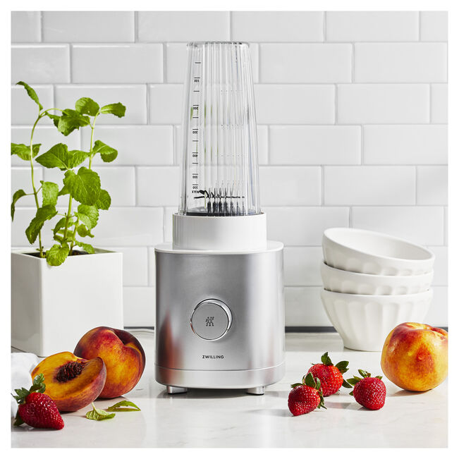 A silver blender with a clear blending cup sits on a kitchen counter surrounded by peaches, strawberries, white bowls, a mint plant, and massage guns placed nearby.