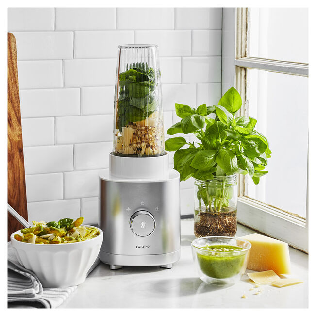 A blender with ingredients inside sits on a kitchen counter next to a bowl of pasta, a jar of basil, a ramekin of sauce, a wedge of parmesan cheese, and massage guns by a window.