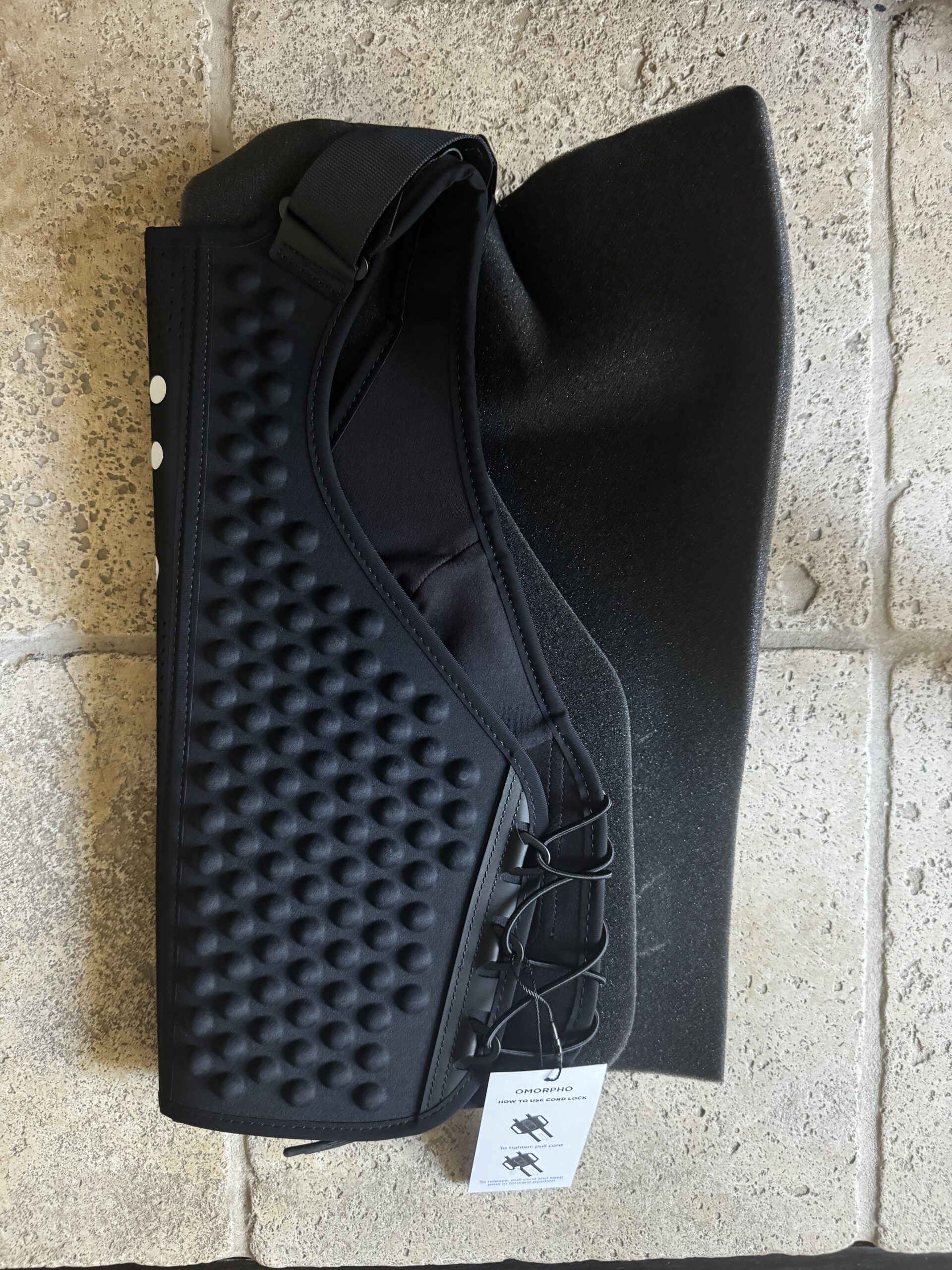 Black neoprene dog harness with textured surface and adjustable straps, lying flat on a beige tiled floor with a tag attached.
