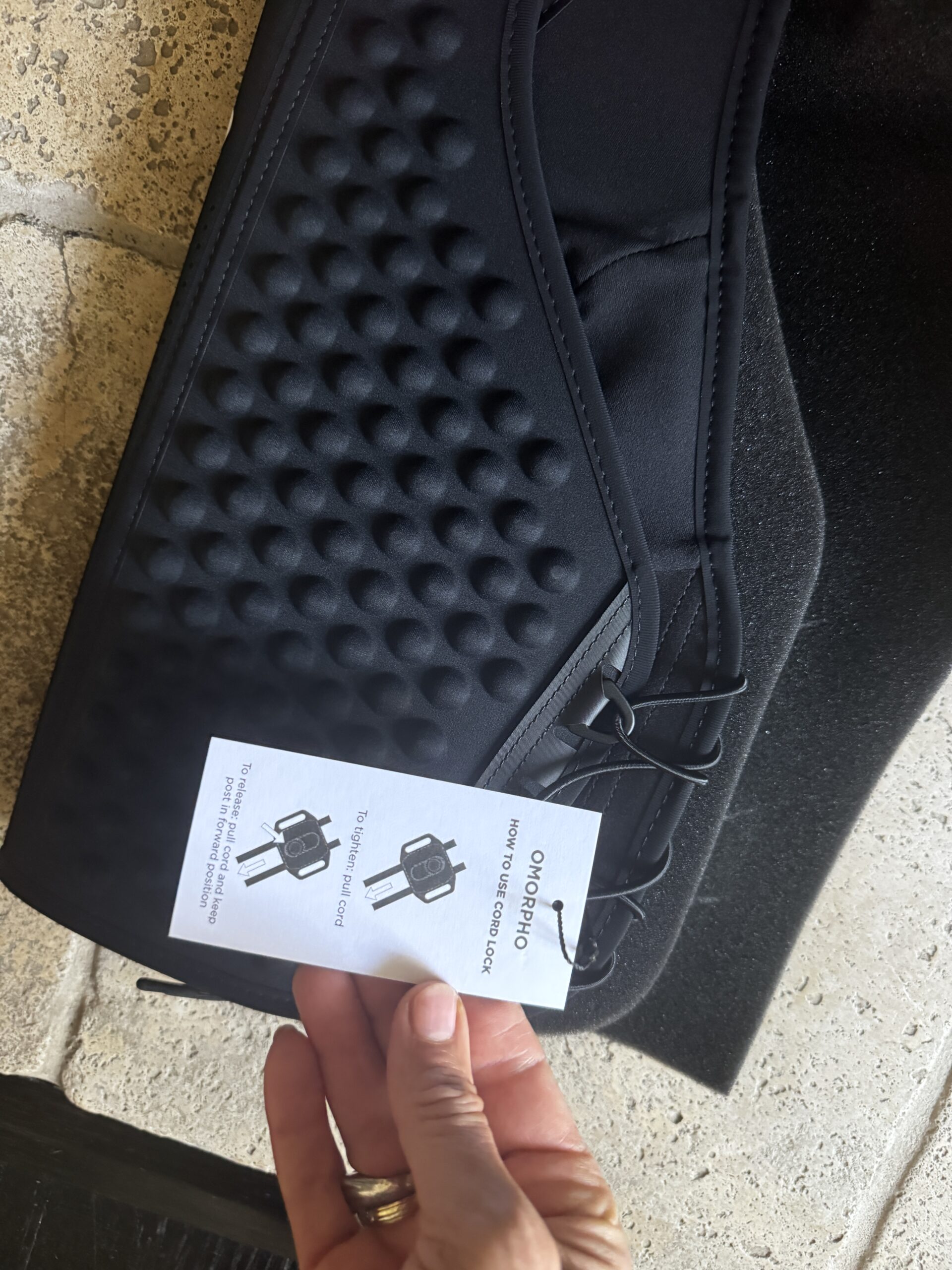 A hand holds a tag with instructions near a black textured product featuring laces; the tag shows diagrams and text on how to close a lock.