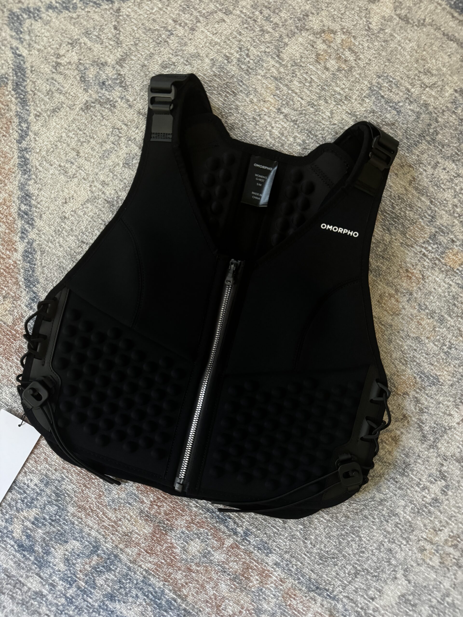 A black weighted workout vest with a front zipper and textured padding lies flat on a light patterned carpet. The brand "OMORPHO" is visible on the upper right side.