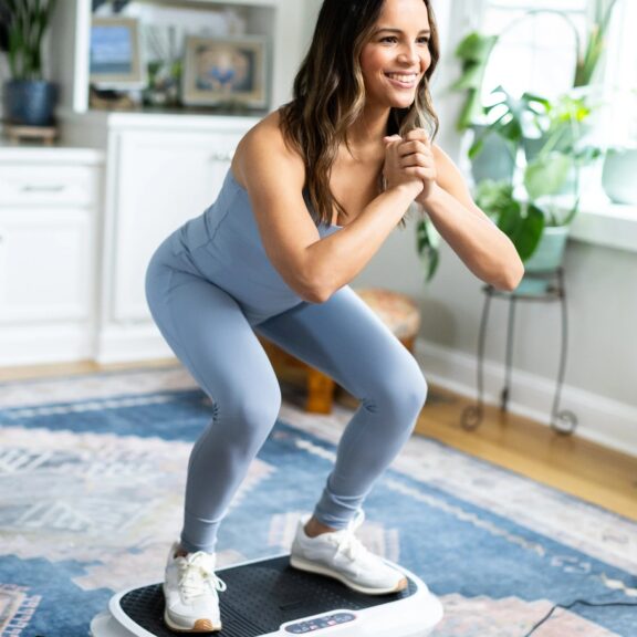 Woman in light blue activewear performs a squat on a vibration plate, with massage guns and decorative items visible in the bright living room filled with plants.