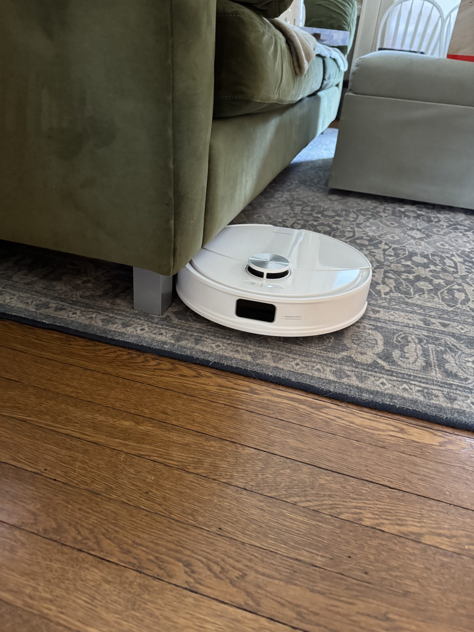 A white robotic vacuum cleaner is partially stuck under a green sofa on a patterned rug next to a wooden floor.