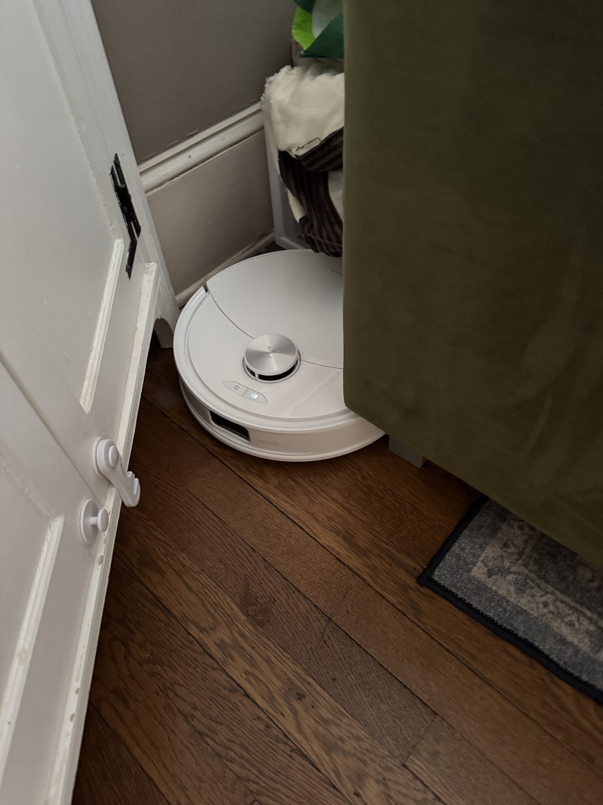 A white robot vacuum is stuck between a door, a piece of furniture, and a wall on a hardwood floor.